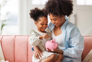 A mother teaching her young daughter about money using a piggy bank.